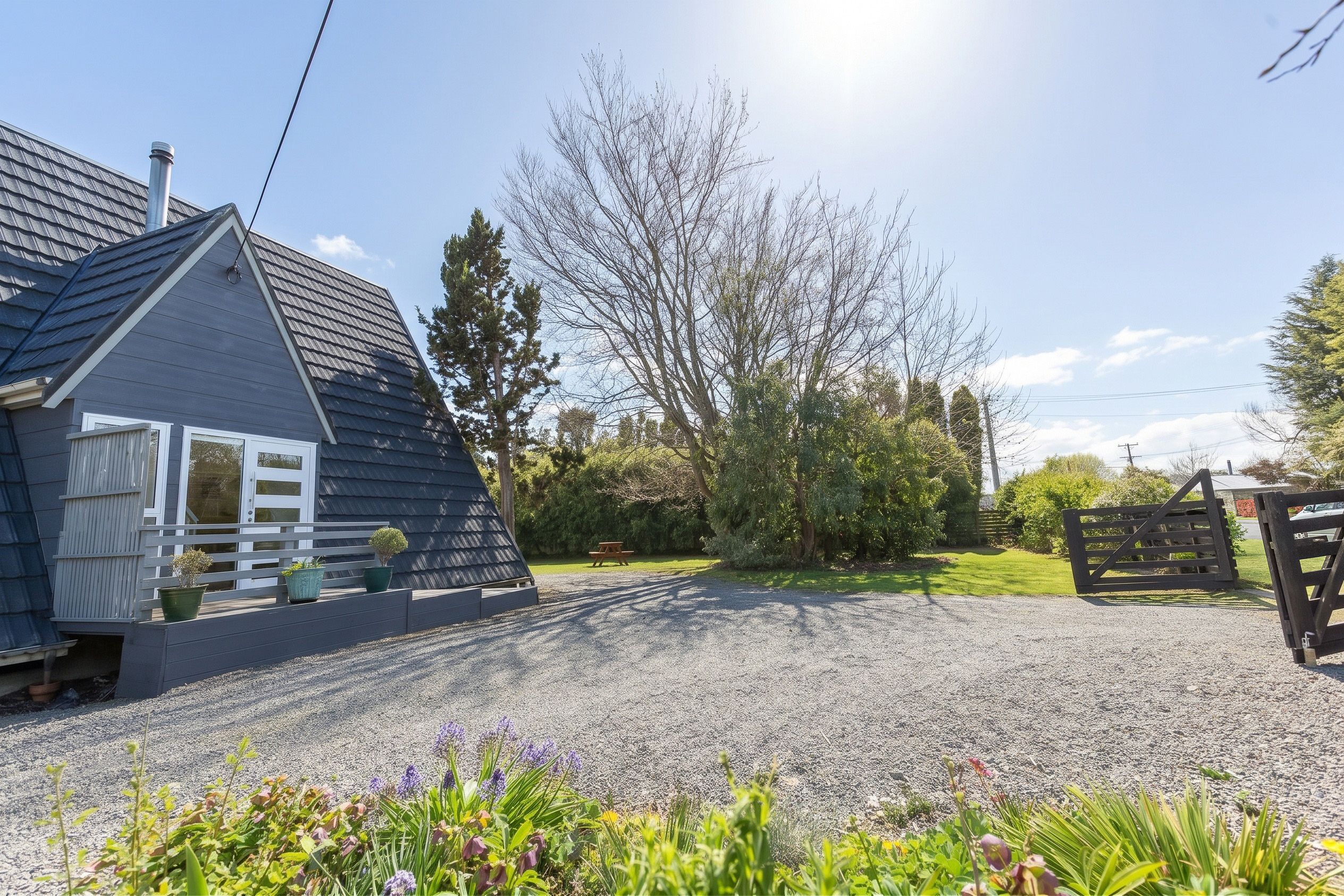 A residential property with a gravel driveway, a wooden gate, and a small house with a sloped roof.