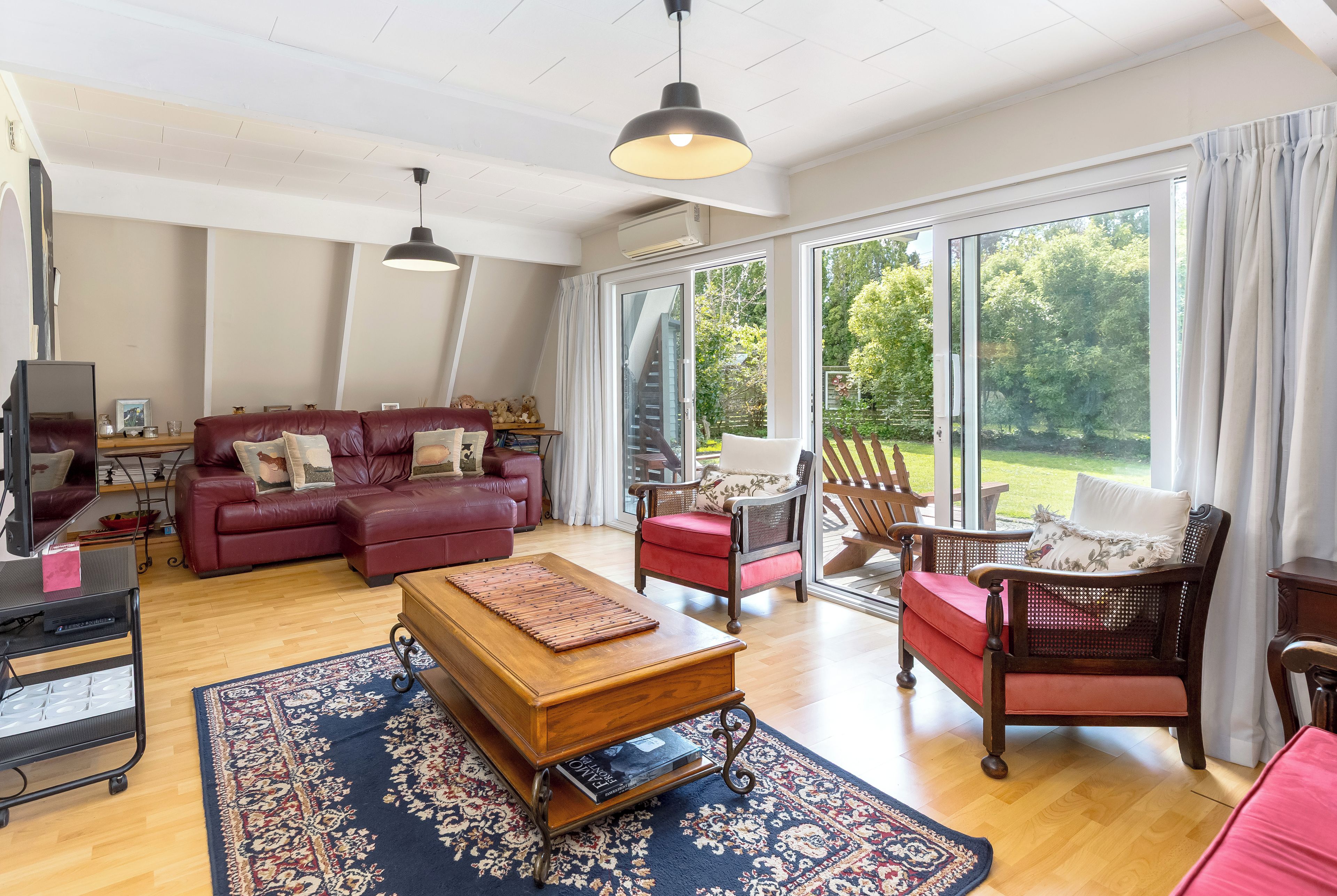 Een ruime woonkamer met een rode bank, een houten salontafel en een rieten fauteuil. Een groot raam biedt uitzicht op het omliggende landschap.