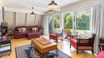 Een ruime woonkamer met een rode bank, een houten salontafel en een rieten fauteuil. Een groot raam biedt uitzicht op het omliggende landschap.
