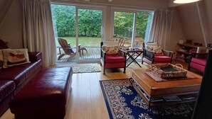 A spacious living room with a red leather sofa, a wooden coffee table, and a large rug. There are two chairs and a small table with a lamp. The room has large windows with curtains, a ceiling fan, and an air conditioning unit. - The house for all seasons, occasions and ages. Perfect for families! (Greytown)