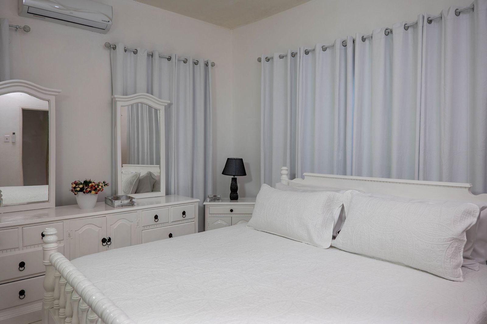 A white bedroom with a large bed, a dresser with drawers, a mirror, a bedside table with a lamp, and a window with curtains.