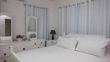A white bedroom with a large bed, a dresser with drawers, a mirror, a bedside table with a lamp, and a window with curtains.