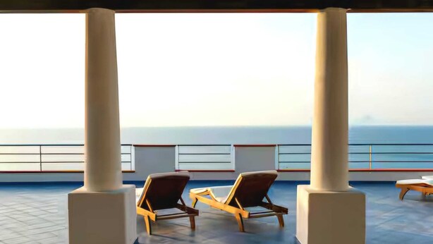 Ein sonniger Außenbereich mit Sitzgelegenheit, zwei Liegestühlen und einem Tisch, mit Blick auf das ruhige Meer.