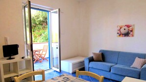 Ein Zimmer mit einem blauen Sofa, einem weißen Tisch mit gemustertem Tischläufer, einem Fernseher auf einem Schrank und einer Glastür zu einem Außenbereich mit Tisch und Stühlen.