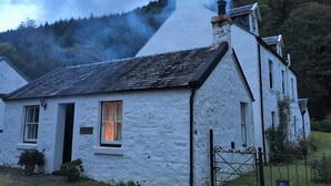 A white cottage with a steep roof, a chimney emitting smoke, and a gate in the foreground. - Quiet village location in beautiful valley close to Dunoon, Argyll. Sleeps 2, pet friendly (Strachur)