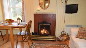 A cozy living room with a fireplace, a dining table with chairs, a television mounted on the wall, and a patterned rug on the floor. - Quiet village location in beautiful valley close to Dunoon, Argyll. Sleeps 2, pet friendly (Strachur)