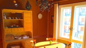 Un coin repas chaleureux avec une table ronde et des chaises en bois, un buffet en bois contenant de la vaisselle, un lustre et une grande fenêtre donnant sur des immeubles.