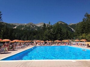 Una piscina con lettini e ombrelloni, circondata da montagne e alberi.