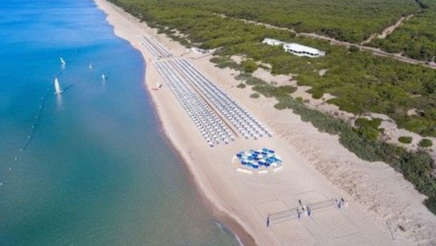 Luchtbeeld van een strand met rijen ligbedden en parasols, een zandstrand en een heldere blauwe zee.