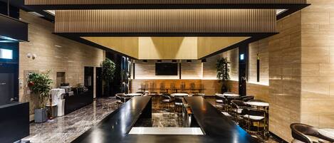 A modern hotel lobby with a long reception desk, seating area, and a large TV screen.