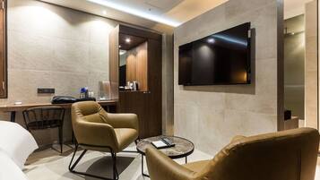 A modern hotel room with a flat-screen TV mounted on the wall, a desk with a chair, and two leather armchairs.
