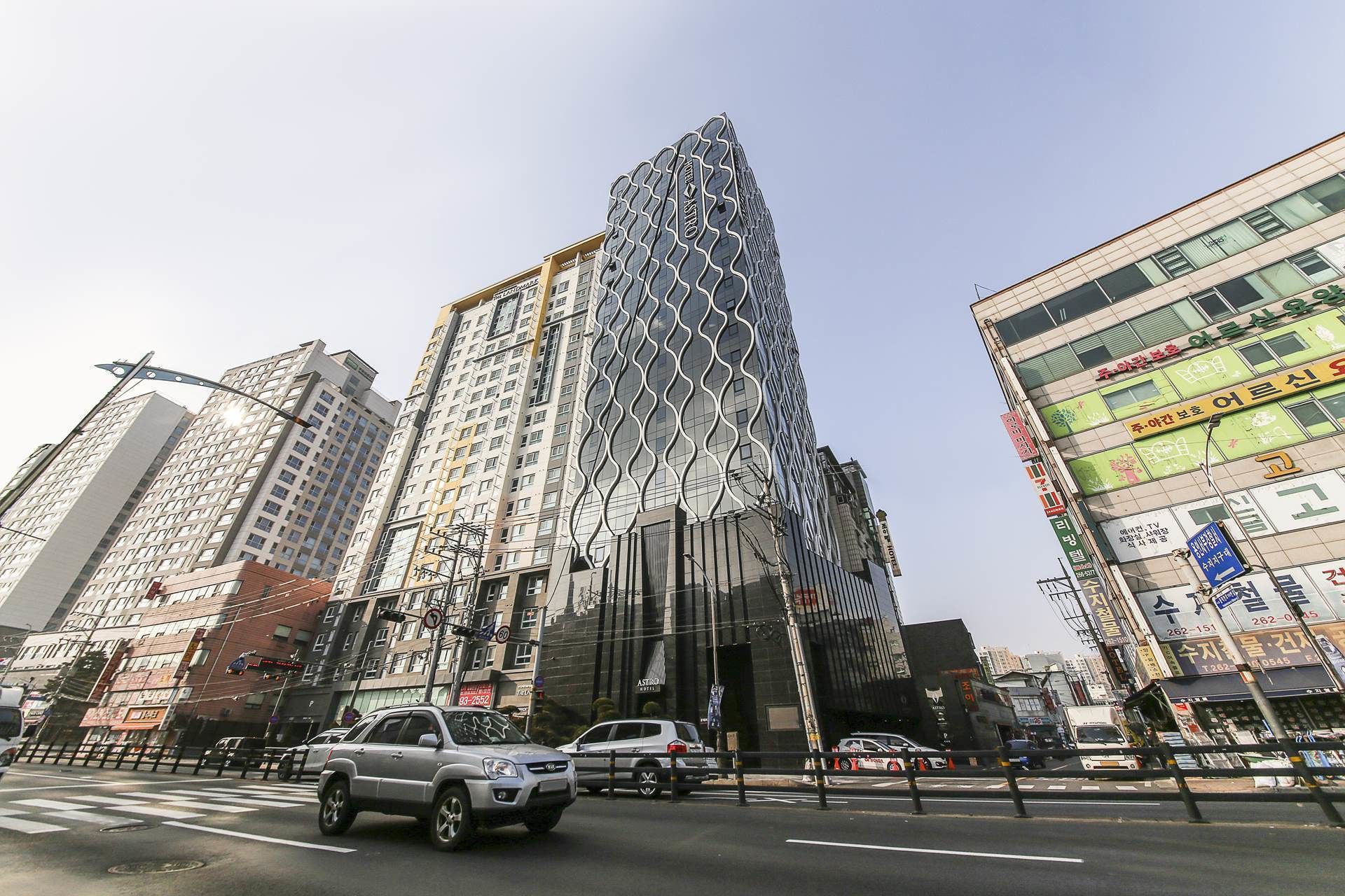 Un edificio moderno de gran altura con una fachada geométrica distintiva, rodeado de otros edificios comerciales y tráfico de calles ajetreado.