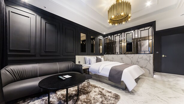 A modern bedroom with a black leather sofa, a round black coffee table, a fluffy rug, a bed with white bedding, and a large mirror. - Yongin Astro Hotel (Yongin)