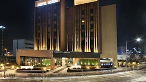 Un hôtel moderne, reconnaissable à son enseigne bien visible où l’on peut lire « HÔTEL ASSEM ». Il se distingue par son grand hall d’entrée illuminé et sa façade bien éclairée.