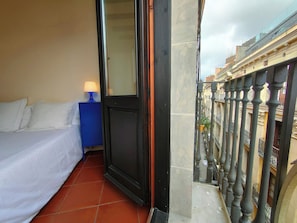 A hotel room with a bed, a nightstand with a lamp, a door, and a view of the cityscape through a balcony railing.