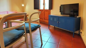 Habitación con televisor en un mueble, una silla con cojín y una ventana con persianas abiertas.