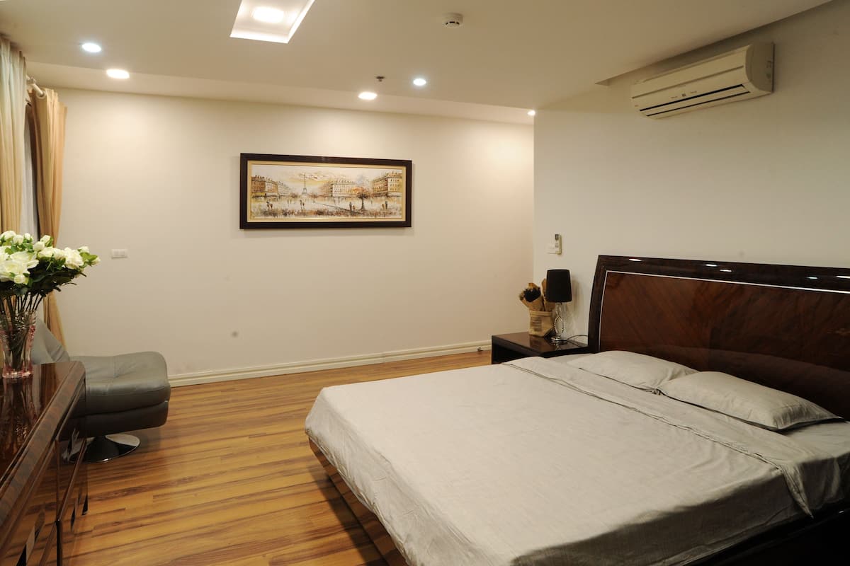 A bedroom with a wooden floor, a bed with a headboard, a nightstand, a lamp, and a framed picture on the wall.