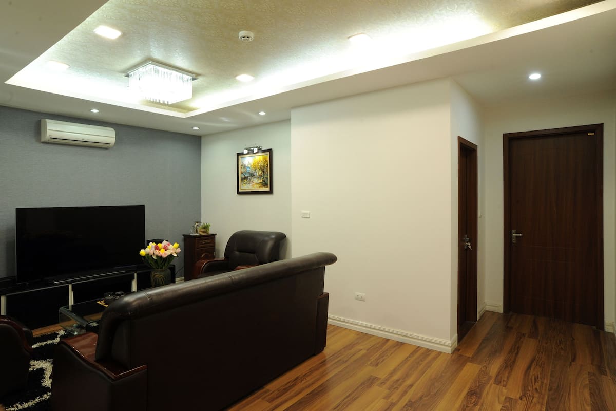 A living room with a flat-screen TV, a dark leather sofa, a wooden floor, and a ceiling with recessed lighting.