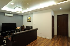 A living room with a flat-screen TV, a dark leather sofa, a wooden floor, and a ceiling with recessed lighting. - Hera Building (Hanoi)