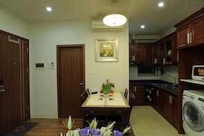 A compact kitchen with wooden cabinets, a built-in washing machine, and a dining area with a table and chairs. - Hera Building (Hanoi)