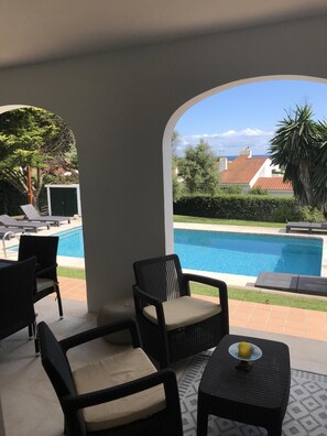 A covered patio area with wicker furniture, a small table with a yellow object, and a view of a swimming pool and surrounding greenery. - 4 bedrooms Villa with pool, Sea view - 3 min walk to Binibeca vell (Binibequer)