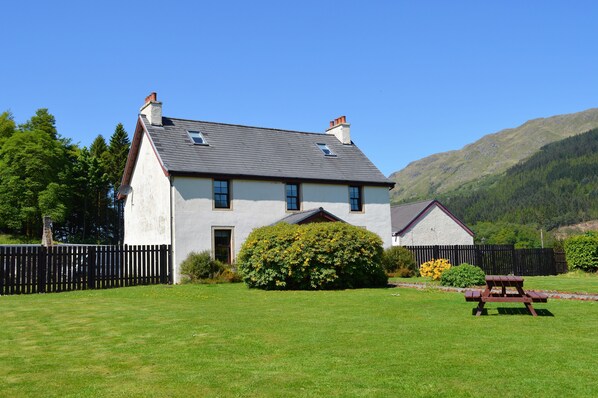 A two-story house with a white exterior, a grey roof, and a small front porch. There is a wooden fence surrounding the property, and a picnic table is visible in the front yard. - Luxury property sleeping 14 with extensive garden, sauna and hot-tub near Dunoon (Ardentinny)
