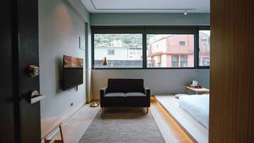 A modern hotel room with a large window, a sofa, a bed, and a small table with a lamp.
