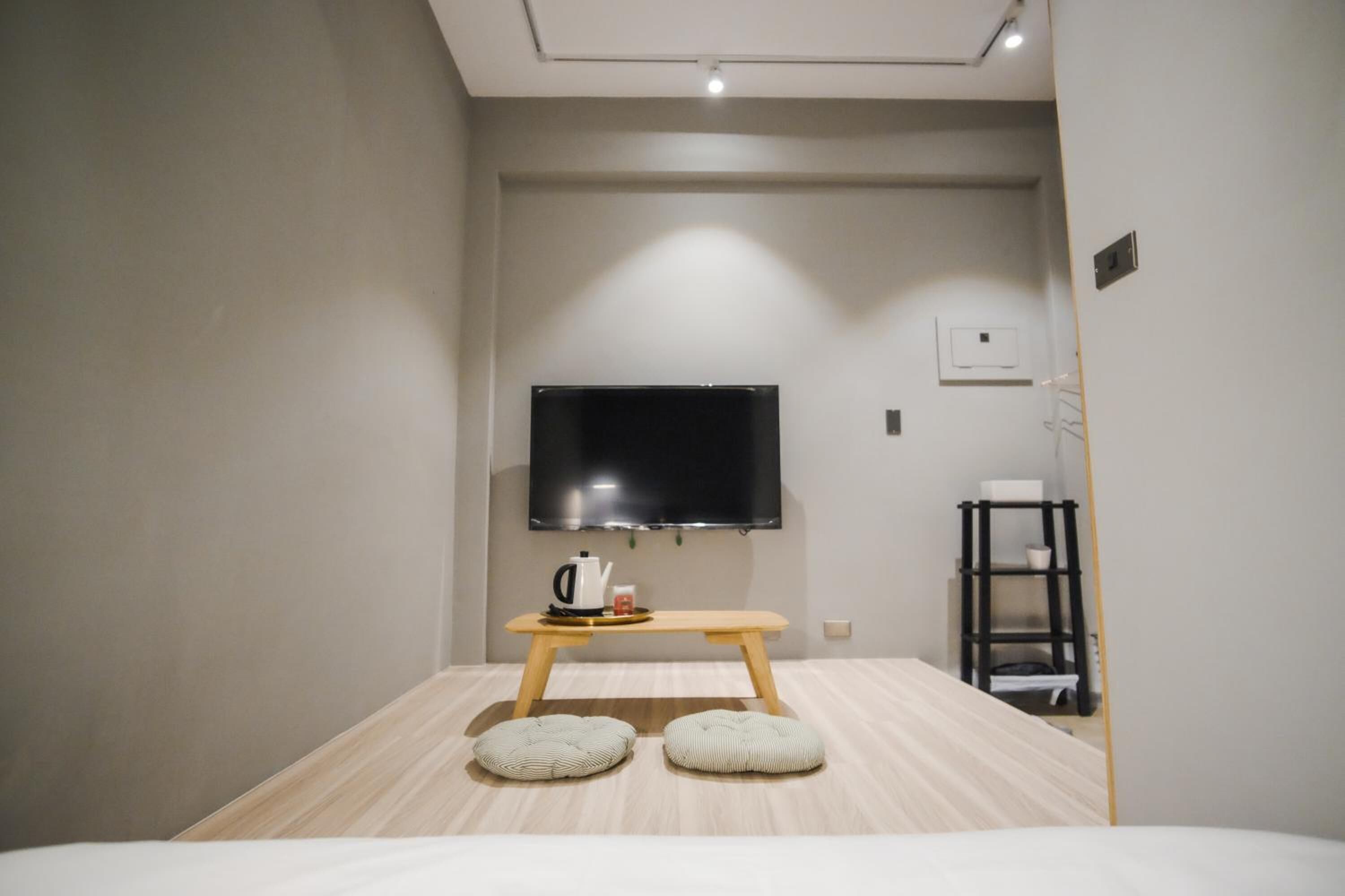 A modern hotel room with a flat-screen TV mounted on the wall, a wooden table with a kettle and cups, two cushioned stools, and a small shelf with items.