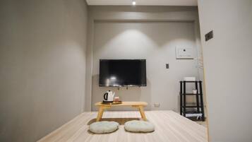 A modern hotel room with a flat-screen TV mounted on the wall, a wooden table with a kettle and cups, two cushioned stools, and a small shelf with items.