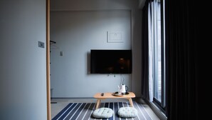 Una habitación moderna con un ventanal grande, un televisor de pantalla plana en la pared, una pequeña mesa de madera con una taza y un plato, y dos taburetes acolchados.