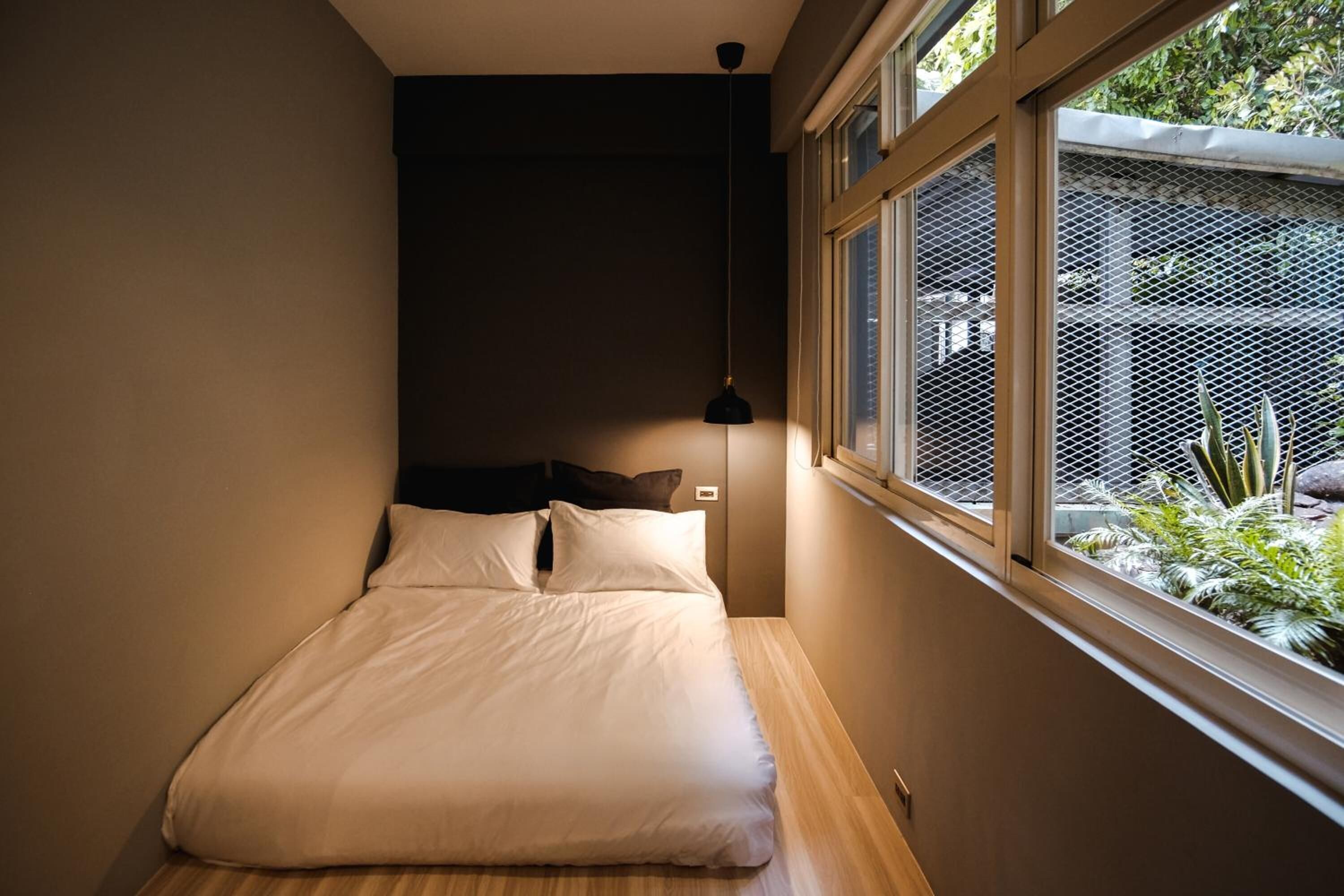 A small, modern bedroom with a bed, a hanging light, and a window with a view of greenery.