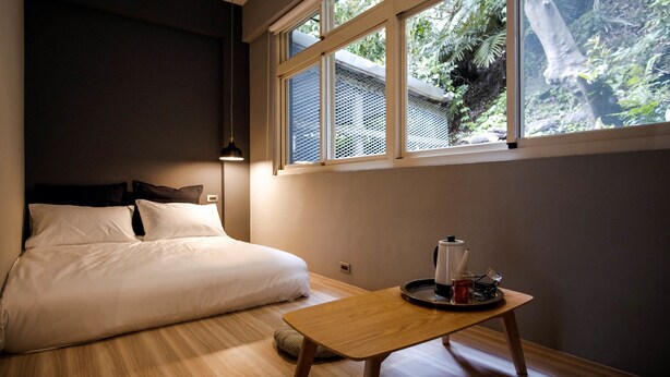 Un dormitorio con cama, una mesita con tetera y ventana con vista a follaje.