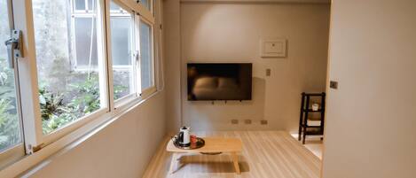 A compact hotel room with a large window, a bed, a small table with a kettle, a TV mounted on the wall, and a wooden bench.