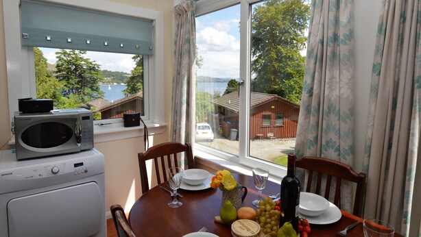 Un coin repas avec une table dressée pour deux personnes, un four à micro-ondes posé sur un lave-linge, et une vue sur un lac et un chalet.