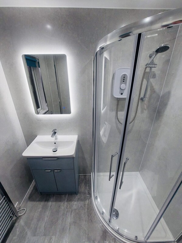 Une salle de bain moderne dotée d’une douche à parois de verre, d’un lavabo blanc et d’un miroir avec éclairage intégré.