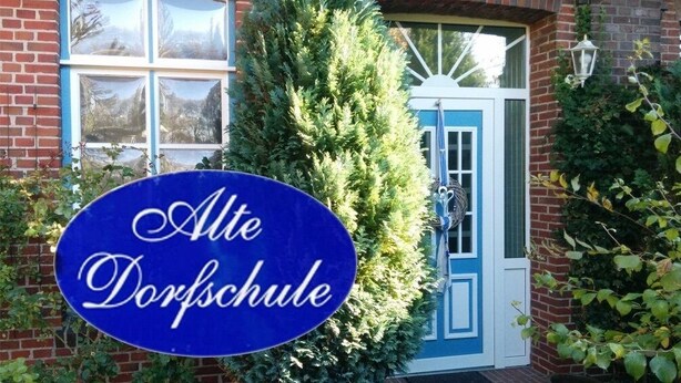 Un panneau bleu sur lequel est inscrit « Alte Dorfschule », devant un bâtiment avec une porte bleue et un mur en briques.