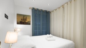 Un lit bien fait, avec du linge de lit blanc, une lampe fixée au mur, un tableau représentant un navire et une fenêtre ornée de rideaux bleus.
