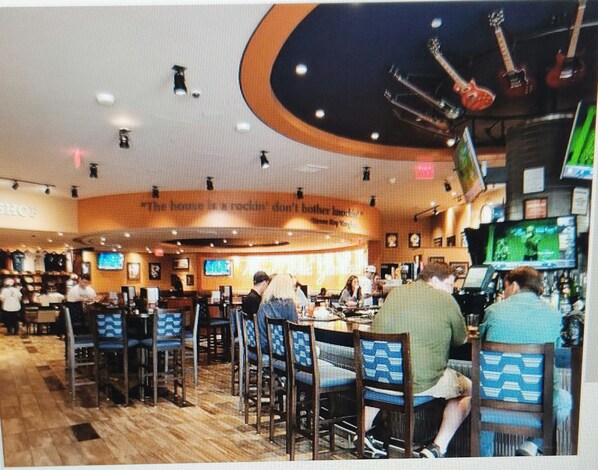 A restaurant with multiple tables and chairs, a bar area with a TV screen, and a ceiling with hanging decorations.