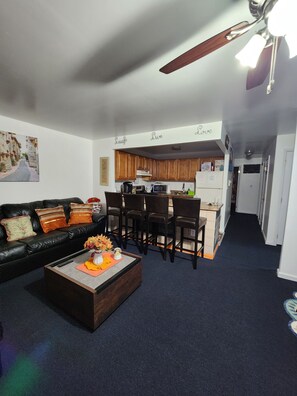 A living room with a black leather sofa, a coffee table with flowers, and a dining area with wooden chairs and a kitchen in the background.