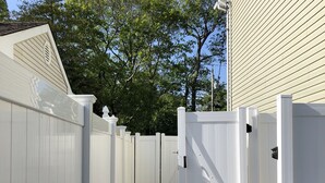 Une clôture en bois peinte en blanc, avec un portail, un chemin de gravier et une maison en arrière-plan.