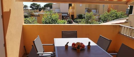 A covered outdoor dining area with a table and chairs, a bowl of fruit, and a view of neighboring buildings and greenery.