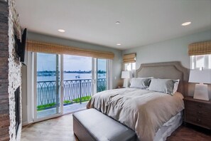 Relax in this serene bedroom with a large bed, bedside tables, and a sliding glass door leading to a balcony with a view of a body of water and cityscape.