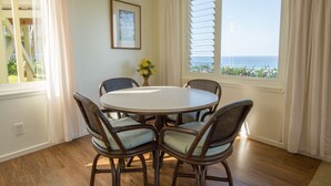Uma sala de jantar com uma mesa redonda e quatro cadeiras de vime, um quadro na parede e vista para o oceano através de grandes janelas.