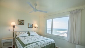 Um quarto com uma cama, duas mesas de cabeceira, um ventilador de teto, uma janela com vista para o oceano e duas luminárias de parede.
