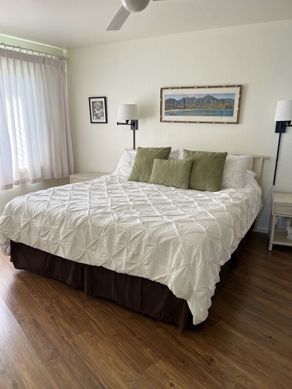 A bedroom with a large bed, wooden flooring, a ceiling fan, and a framed landscape painting on the wall.