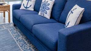 Un canapé bleu agrémenté de coussins aux motifs marins, un tapis à motifs et une vue sur un coin repas avec des chaises et une table.