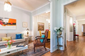 A living room with a sofa, armchairs, a coffee table, and a rug. There is a painting on the wall and a vase with flowers on a side table.