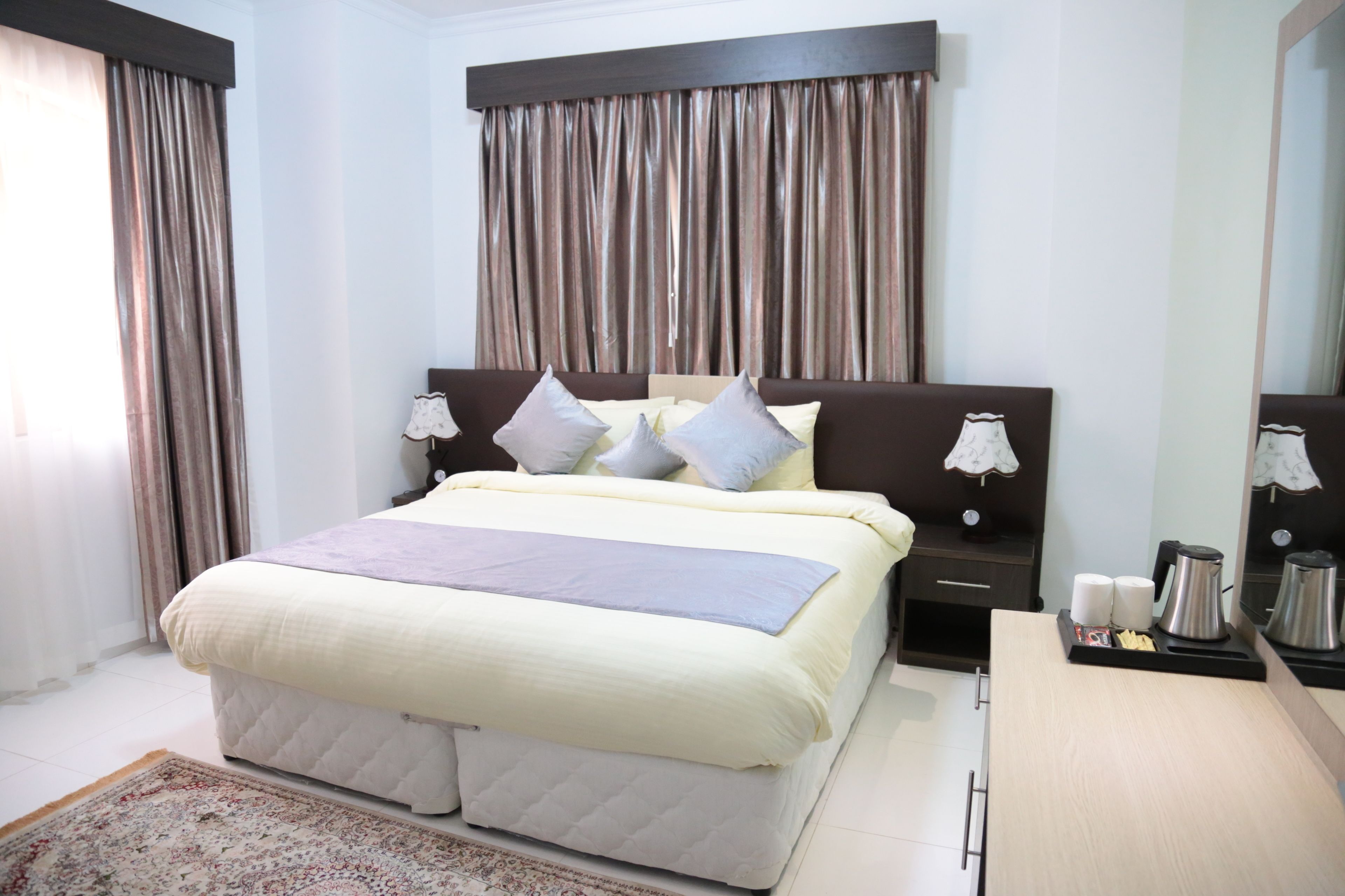 A hotel room with a large bed, a bedside table with a lamp, a nightstand, a wooden desk, and a coffee maker.