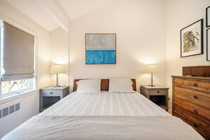 Een slaapkamer met een bed, twee nachtkastjes, een dressoir, een raam met gordijn en een schilderij aan de muur.