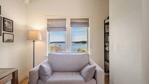 Une pièce avec un canapé gris, une lampe, une bibliothèque et une fenêtre donnant sur la mer.